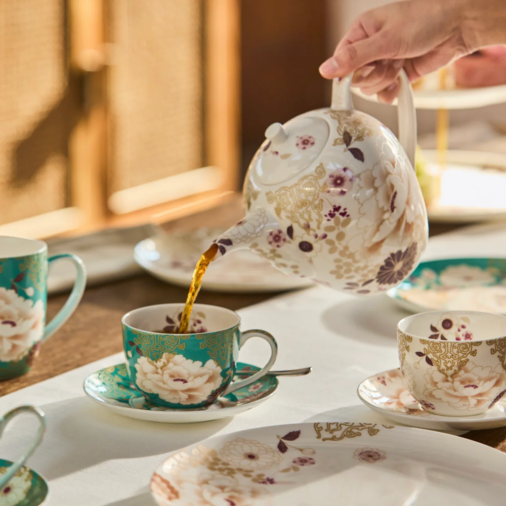 Tea being poured from a floral teapot into a matching cup and saucer on a table.