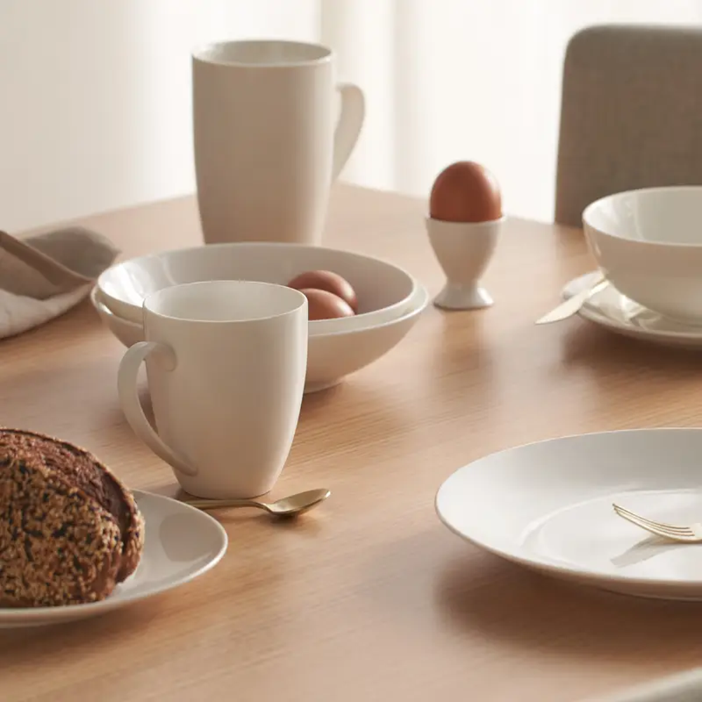 Breakfast setting with white mugs, bowls, and a loaf of bread on a wooden table.