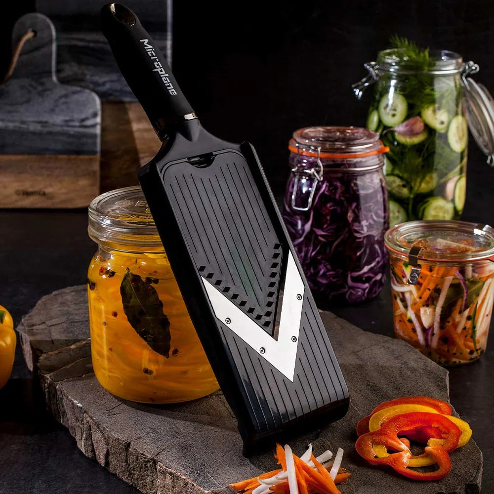 Microplane grater on a wooden board with jars of pickled vegetables in the background
