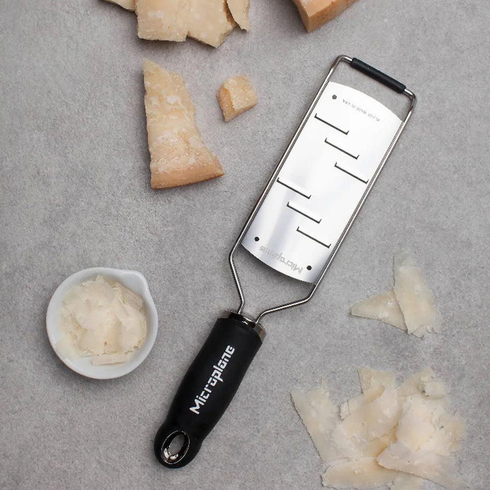 Microplane zester on a gray surface with grated cheese and Parmesan rinds.