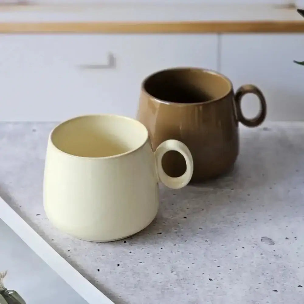 Two ceramic mugs, one beige and one brown, on a speckled countertop.