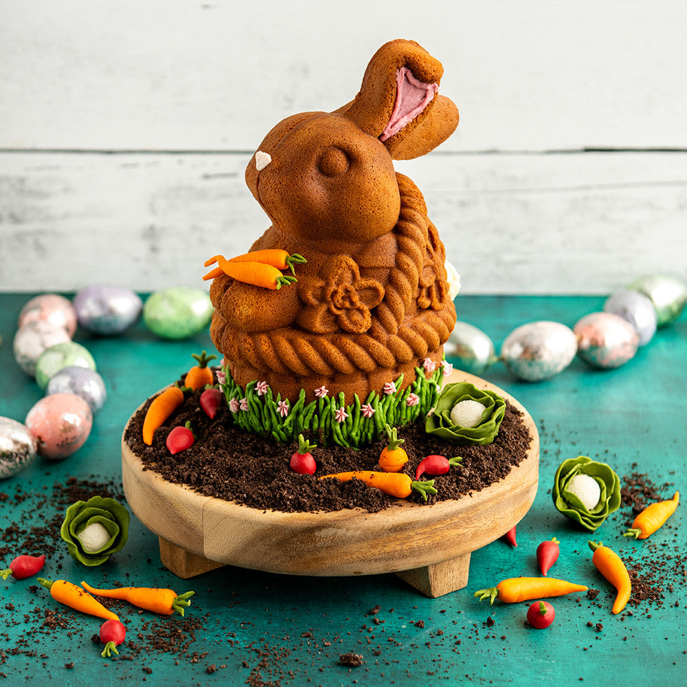 Decorative Easter bunny shaped cake with a wooden base, surrounded by Easter eggs and carrots on a teal surface.