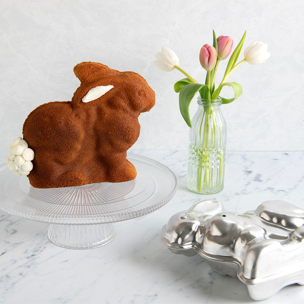 Bunny-shaped cake on a stand with a vase of tulips and a metal egg carton on a marble surface.
