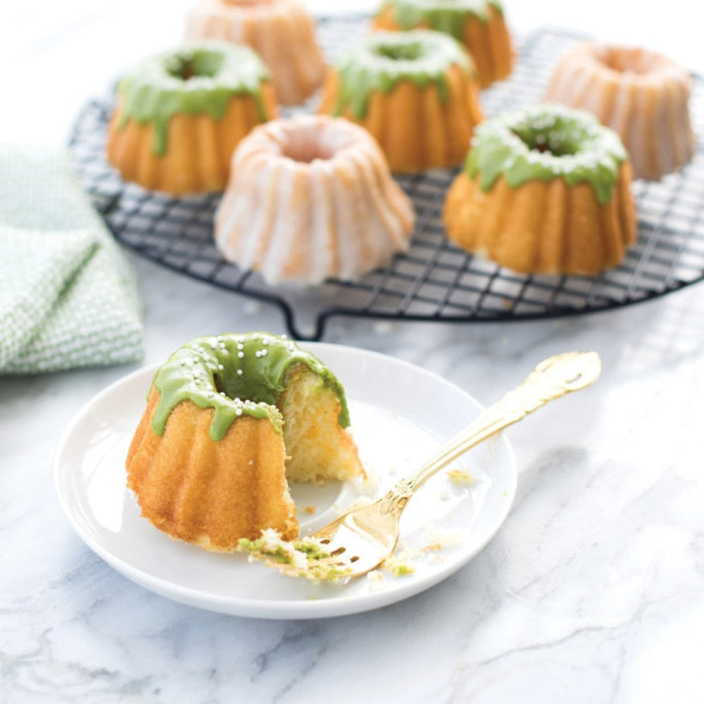Small bundt cakes with green glaze on a white plate with a gold fork, more cakes in the background.