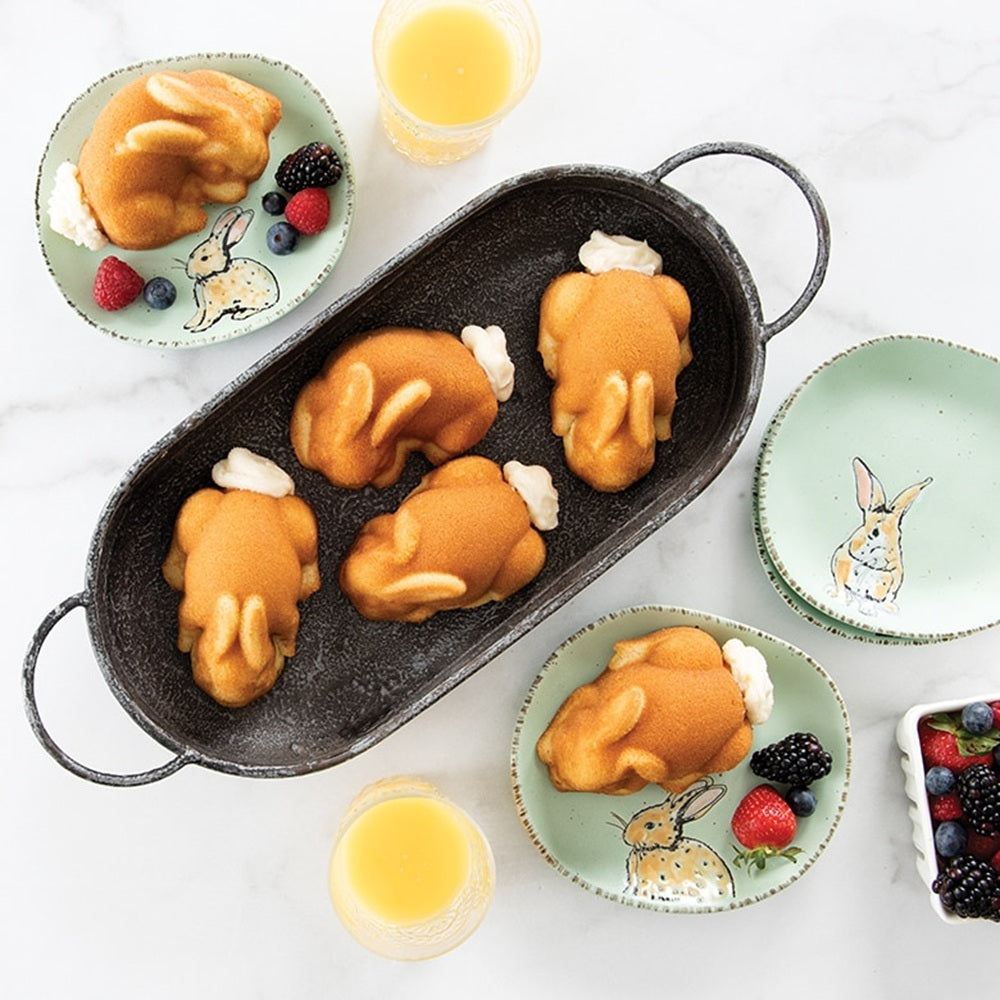 Bunny-shaped pancakes on a pan with small plates and glasses of orange juice on a white background.