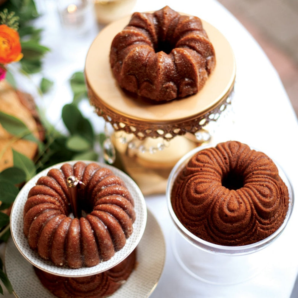 Three small ring-shaped bundt cakes in different baked flavors displayed on stands and plates on a white table, with green leaves and flowers in the background.