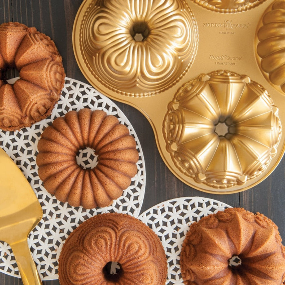 A gold-colored bundt cake pan with decorative floral patterns is shown alongside several baked ring-shaped bundt cakes on white patterned paper mats and a gold spatula, all arranged on a dark wood surface.