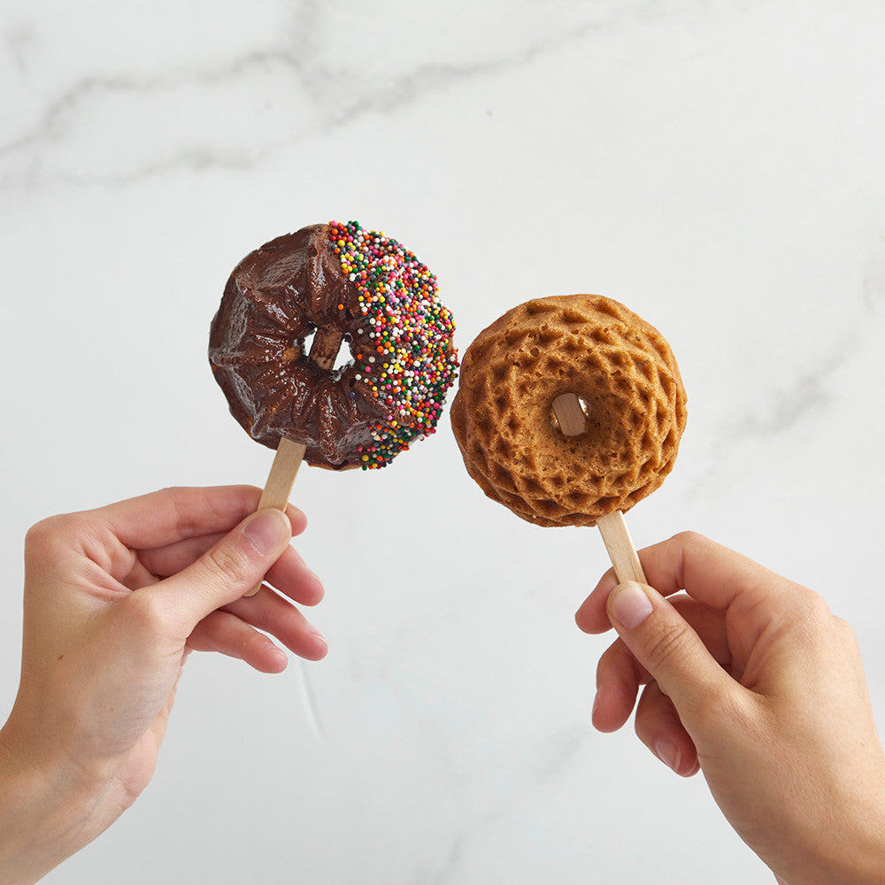 Two hands holding novelty donut-shaped lollipops against a light marble background. One lollipop has a chocolate glaze with rainbow sprinkles, and the other is a textured golden-brown ring donut.