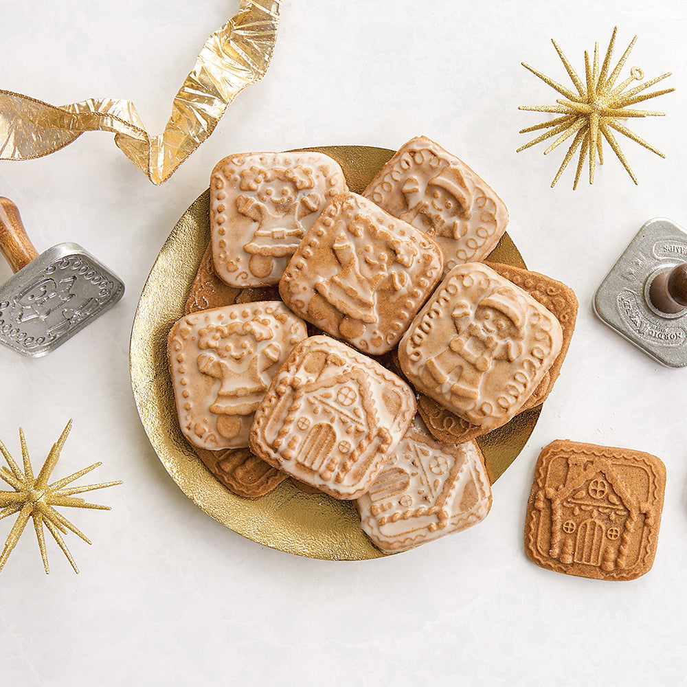 A styled flat-lay of gingerbread cookies with embossed holiday designs, served on a gold plate with gold and silver cookie cutters on a white background.
