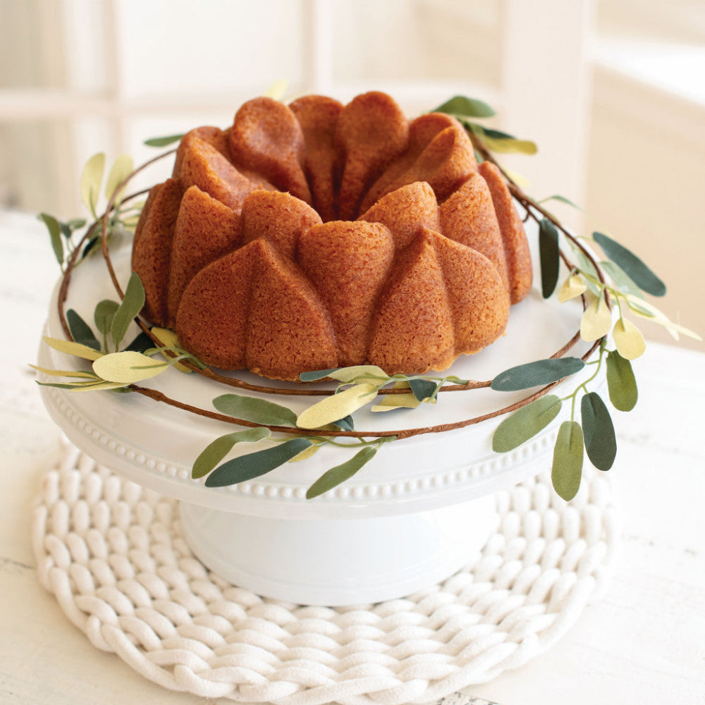 A golden-brown bundt cake displayed on a white pedestal stand, decorated with a green and brown leafy garland, set on a white woven placemat in a bright indoor setting.