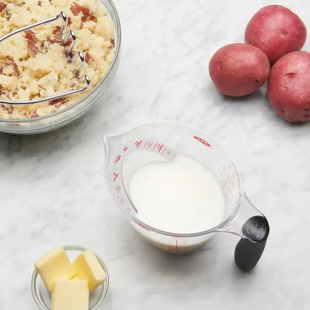 Mashed potatoes in a bowl with a measuring cup of milk, butter, and red potatoes on a marble surface.