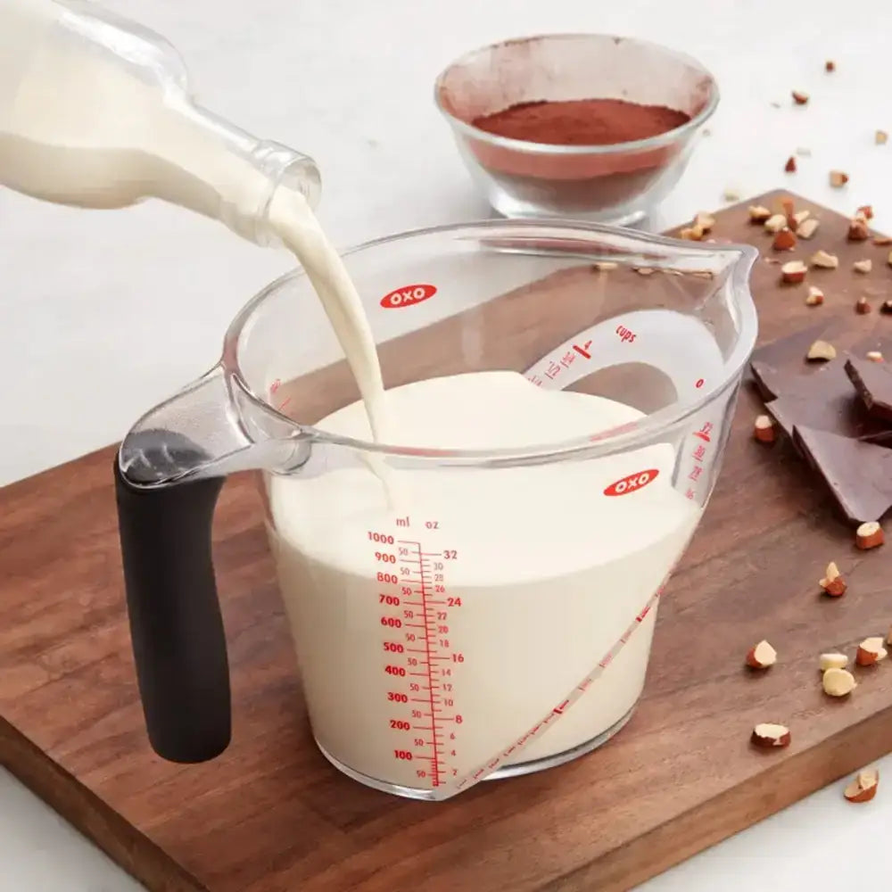 Measuring cup being filled with milk on a wooden cutting board with chocolate and nuts.