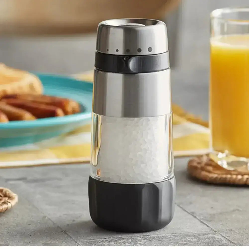 Stainless steel salt grinder on a table with a glass of orange juice and cinnamon sticks in the background.