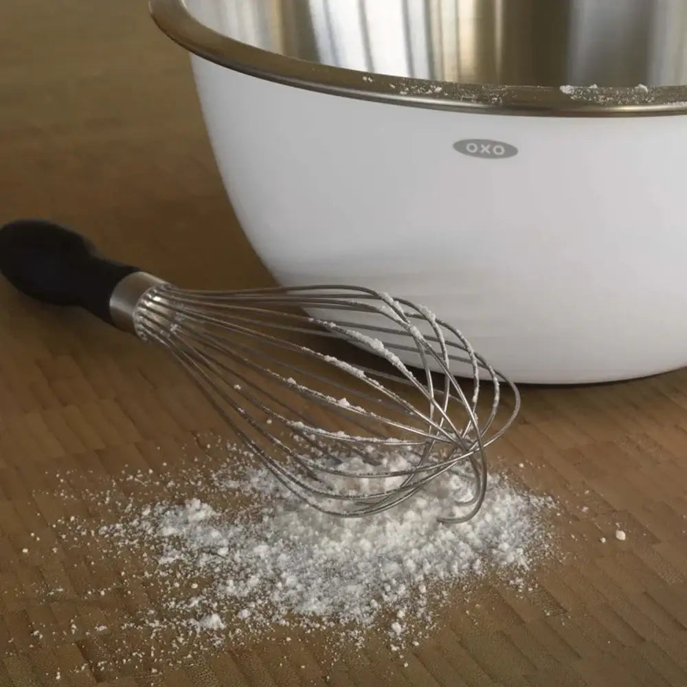 White mixing bowl with black handle and metal whisk on a wooden surface with flour.