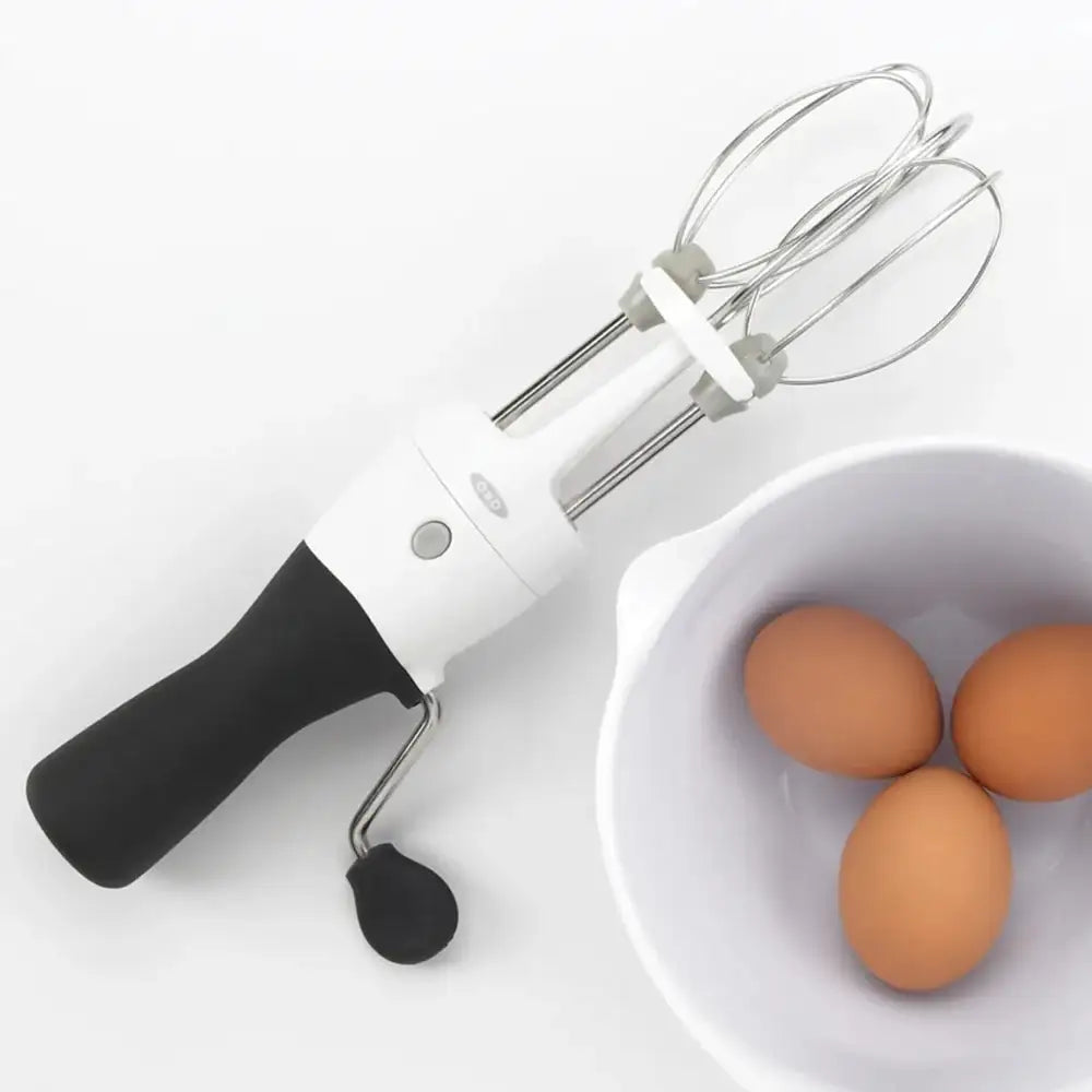 Handheld electric mixer with whisk attachment next to a bowl of eggs on a white background