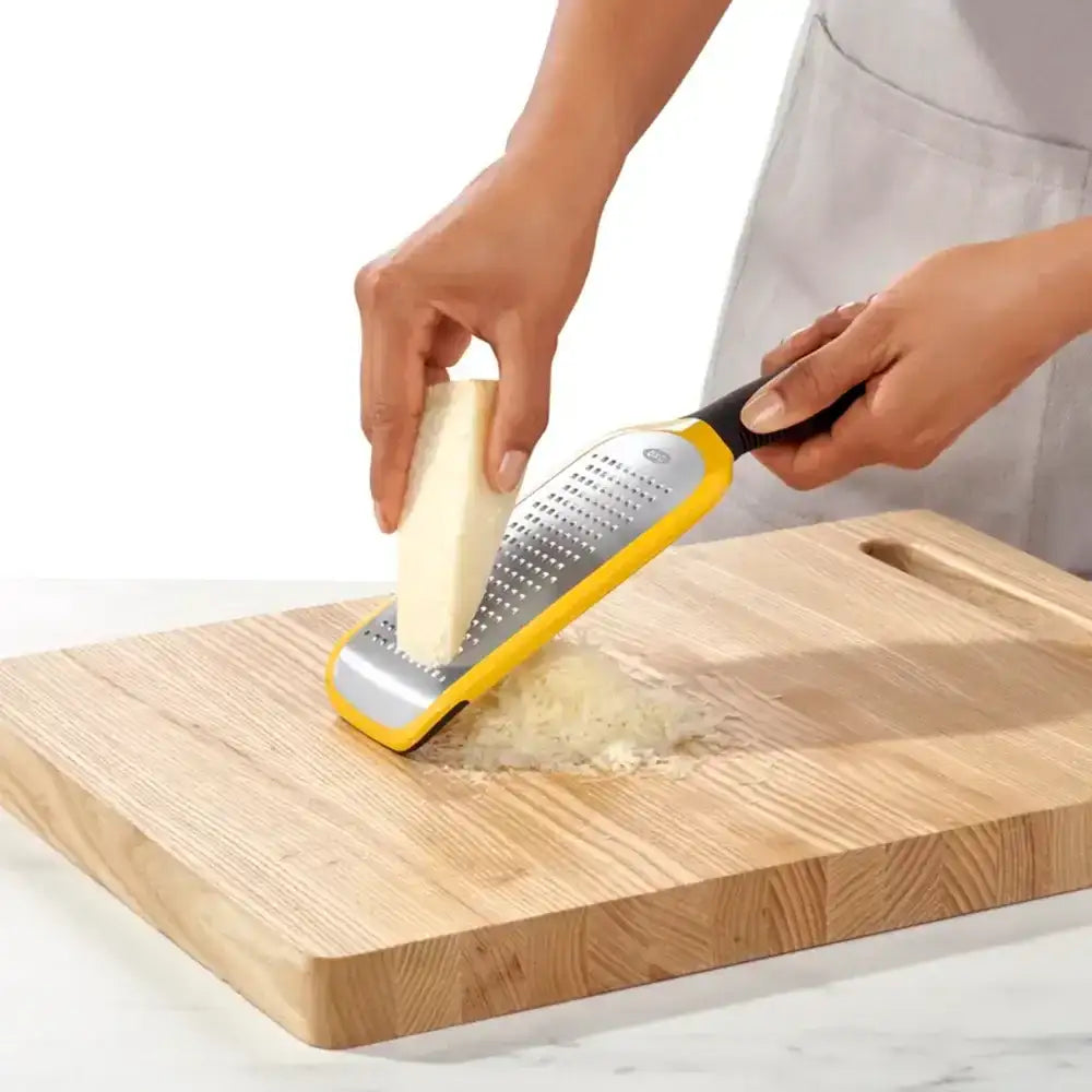 Person using a yellow and silver grater on a piece of cheese over a wooden cutting board.