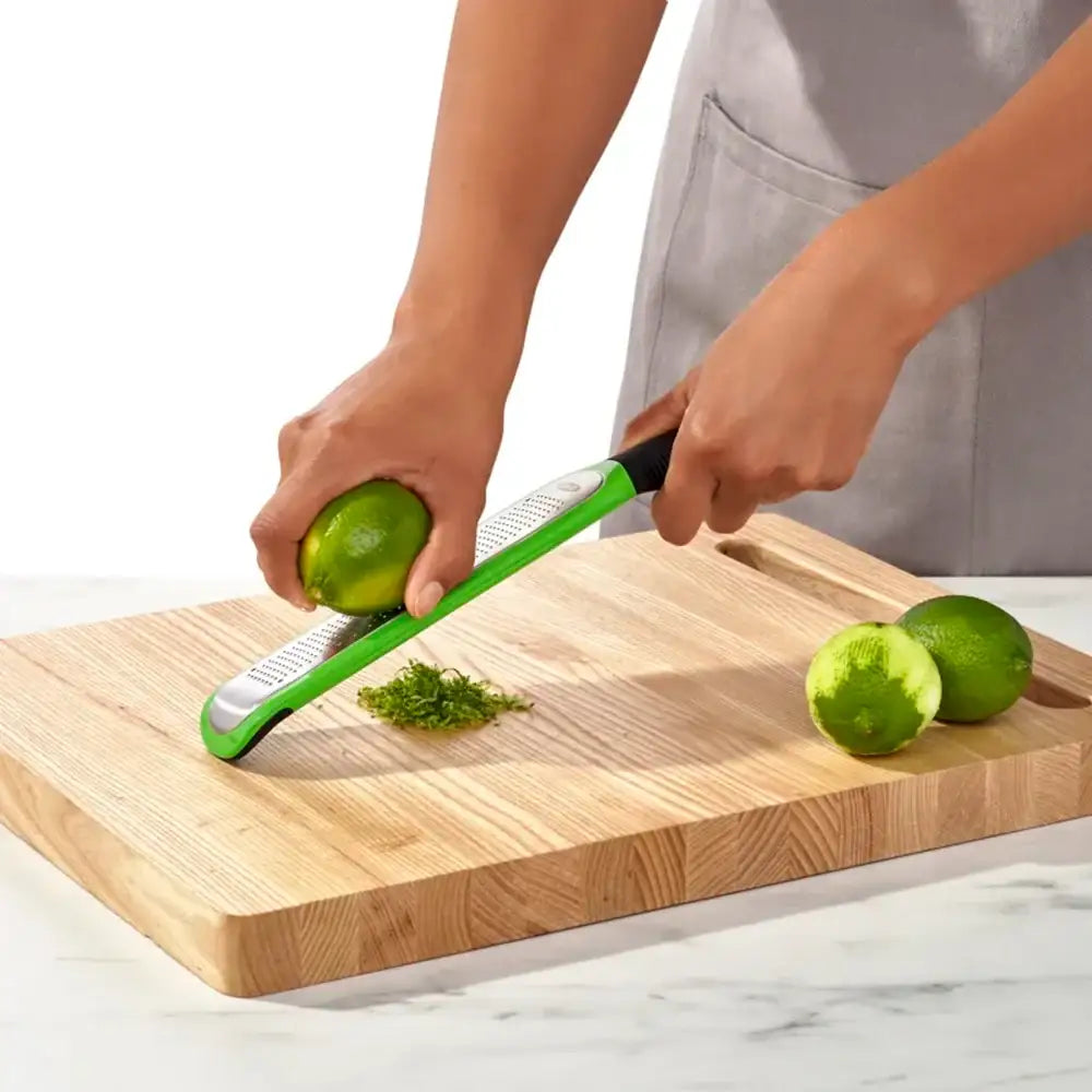 Person using a green citrus zester on a lime on a wooden cutting board.