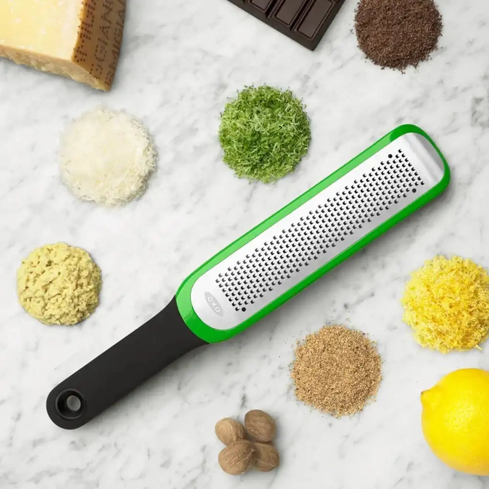 Handheld grater with black handle and green top on a marble surface with various ingredients.