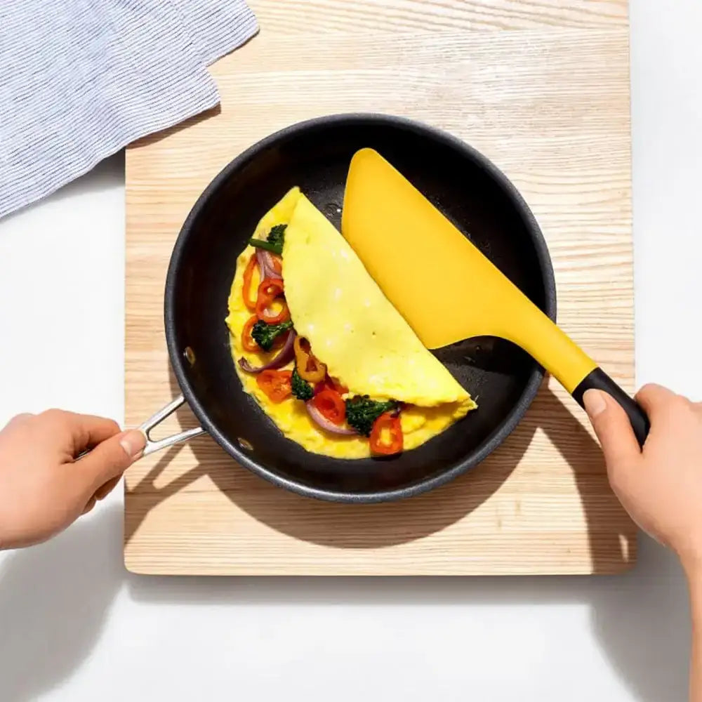 Person flipping an omelette in a pan with a yellow spatula on a wooden cutting board.
