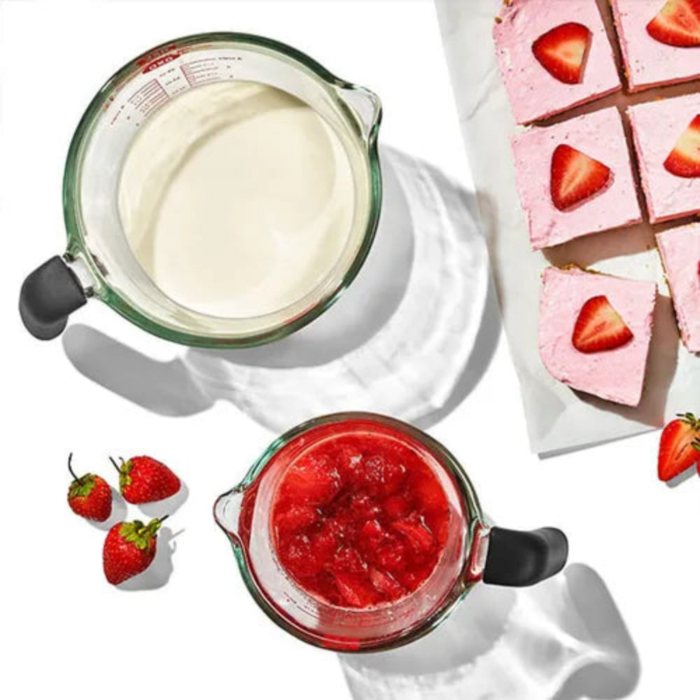 Two glass measuring jugs with strawberry puree and cream, and a sliced strawberry cake on a white surface.