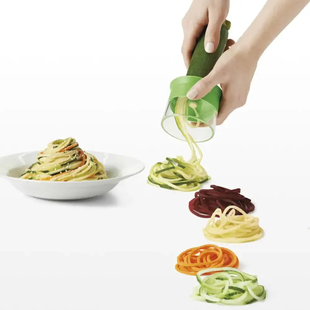 Hand using a green spiralizer to create zucchini noodles with a bowl of prepared noodles on a white background.