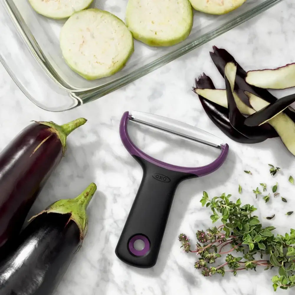 Purple and black vegetable peeler on a marble surface with eggplants and herbs.