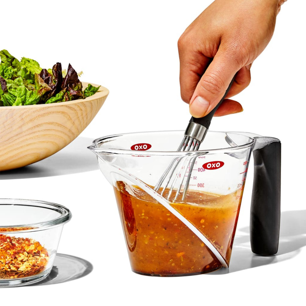 Person mixing salad dressing in a measuring cup with a bowl of greens and spices in the background.