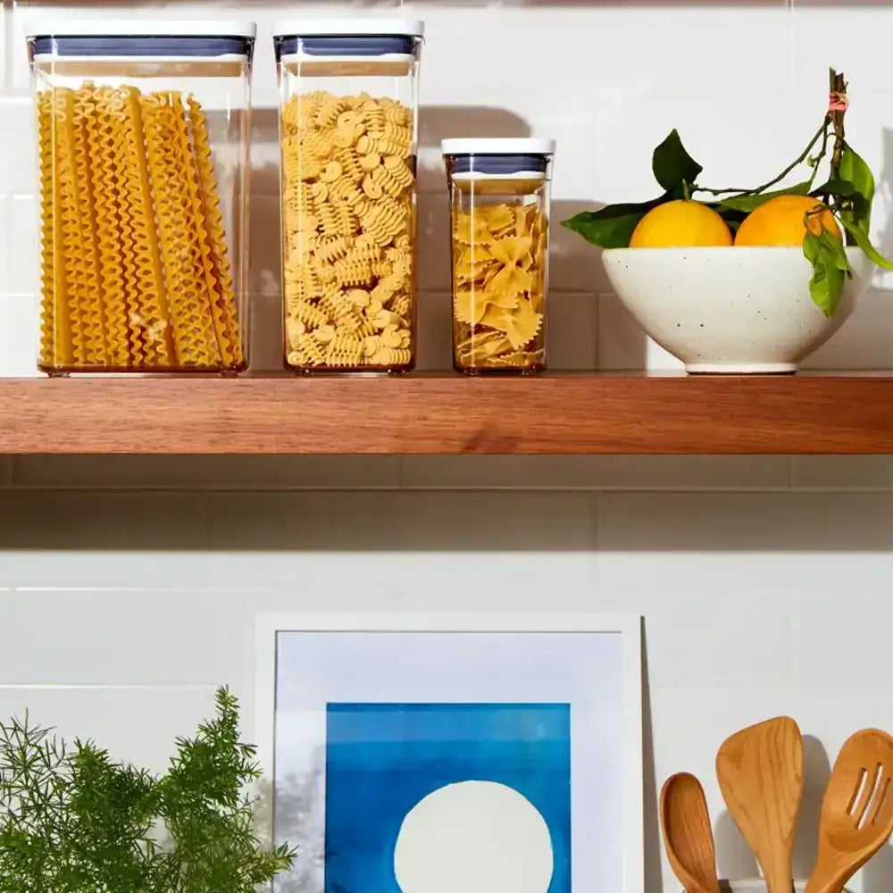 Kitchen shelf with pasta containers, a bowl of fruit, and a framed picture.