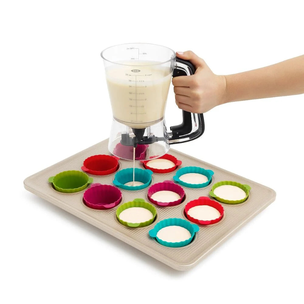 Person dispensing batter into colorful silicone cupcake liners on a baking tray.