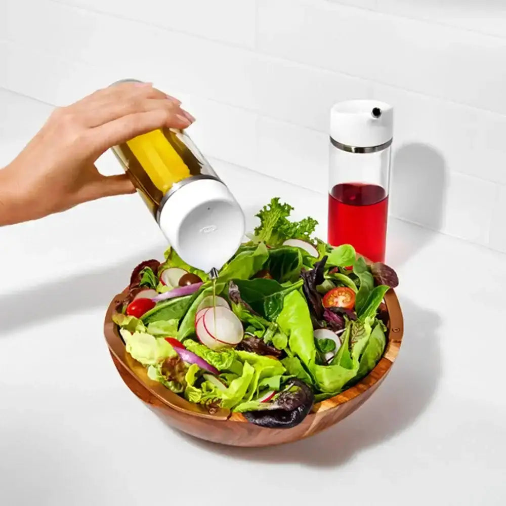Person pouring dressing from a glass bottle onto a salad with a second bottle of dressing in the background.
