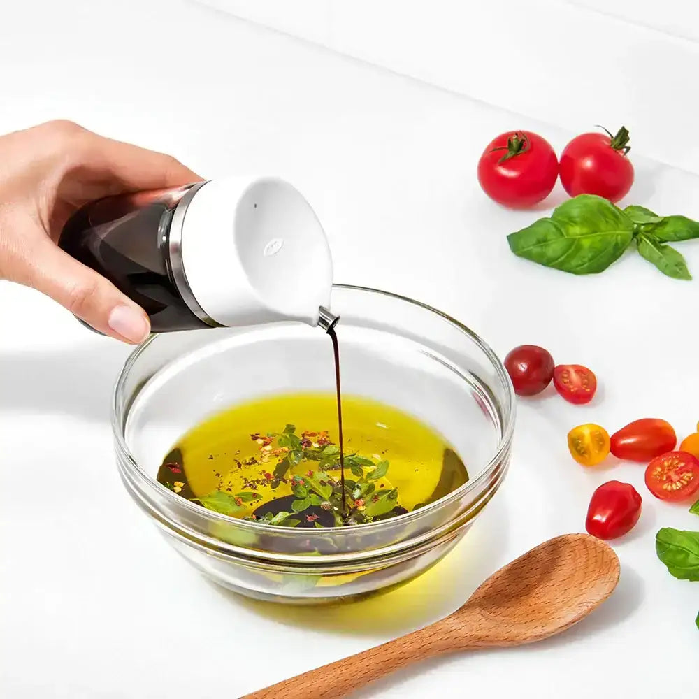 Hand pouring olive oil into a glass bowl with tomatoes and basil on a white background