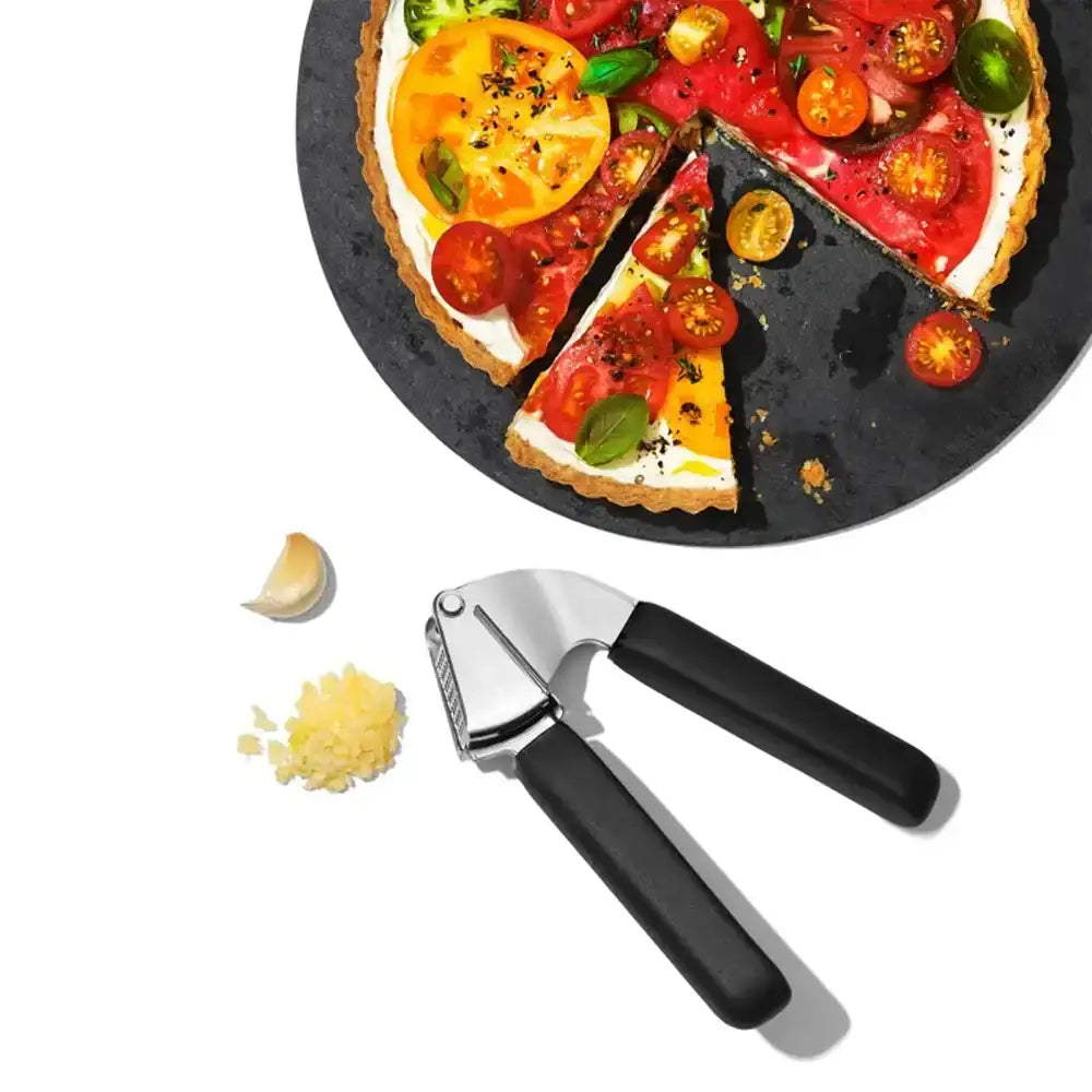 Tomato tart with a slice cut out, garlic press, and garlic on a white background