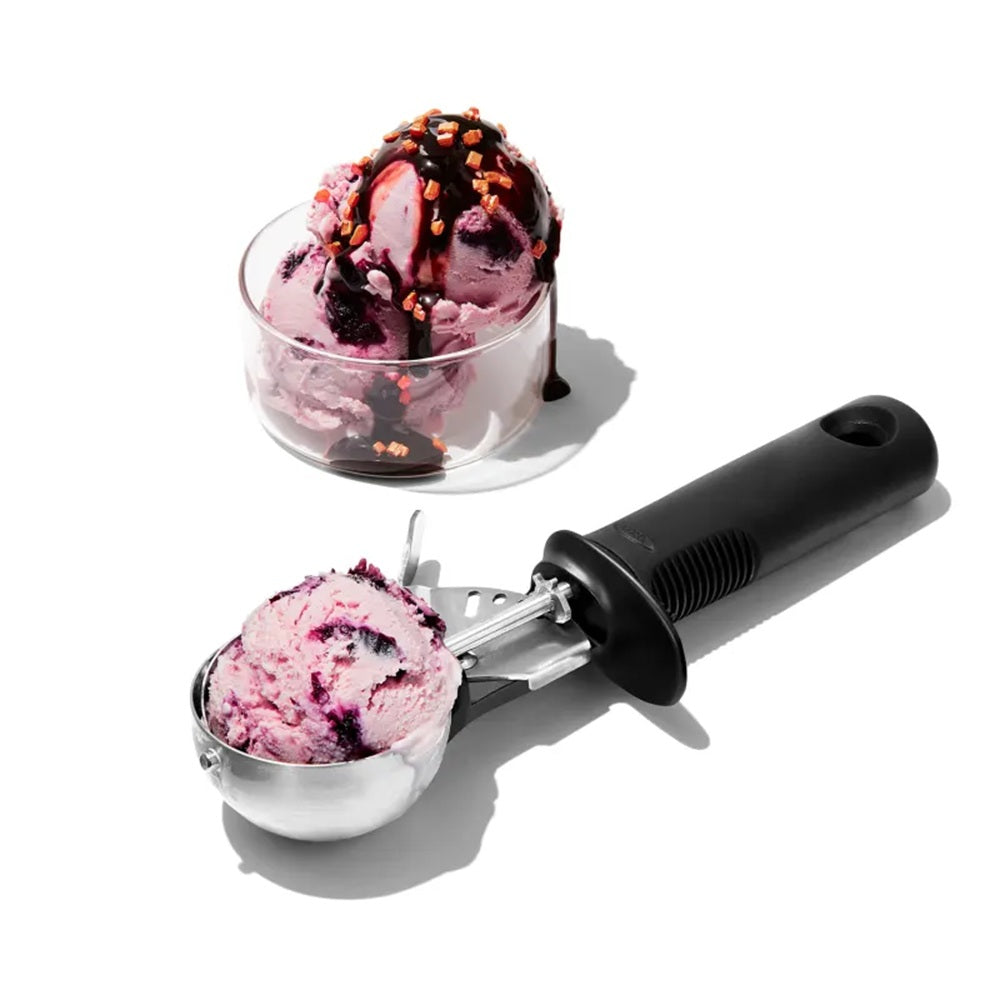 Ice cream scoop with pink ice cream next to a glass bowl of ice cream on a white background