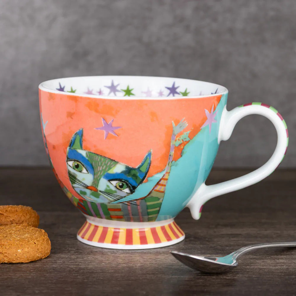 Colorful mug with a cat design on a wooden surface with cookies and a spoon.