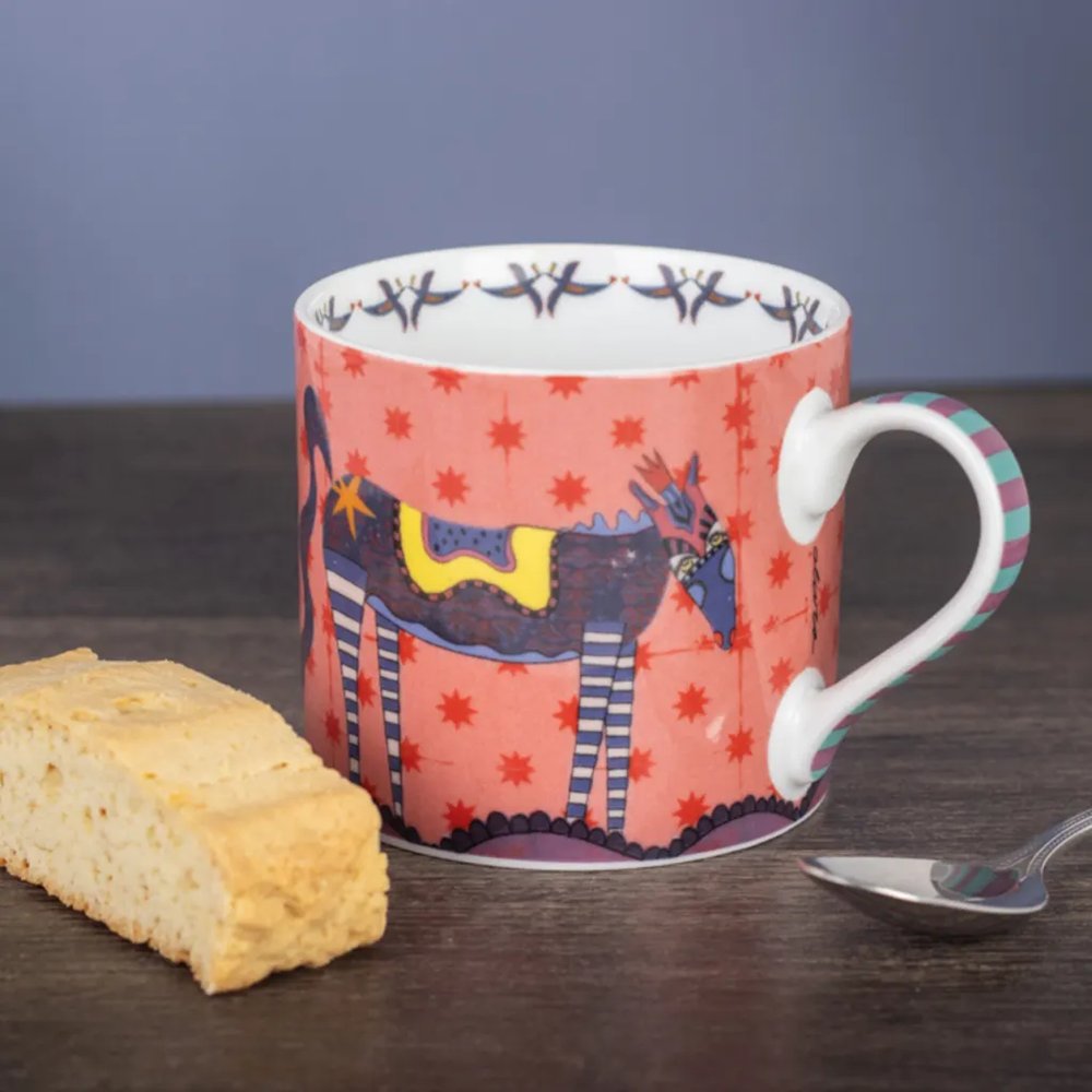 Colorful mug with horse design on a wooden surface with a biscuit and spoon.