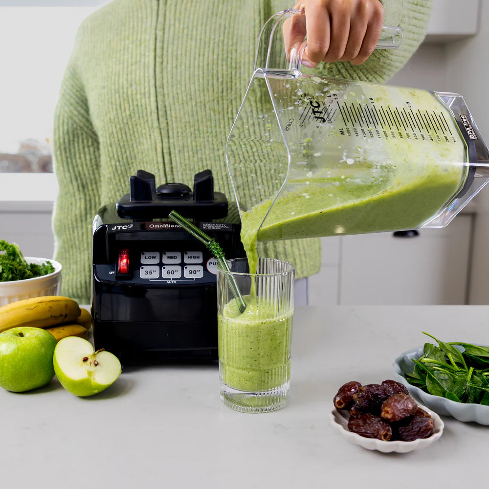 Person pouring a green smoothie from a blender into a glass, with ingredients like bananas, apples, and dates on the counter.
