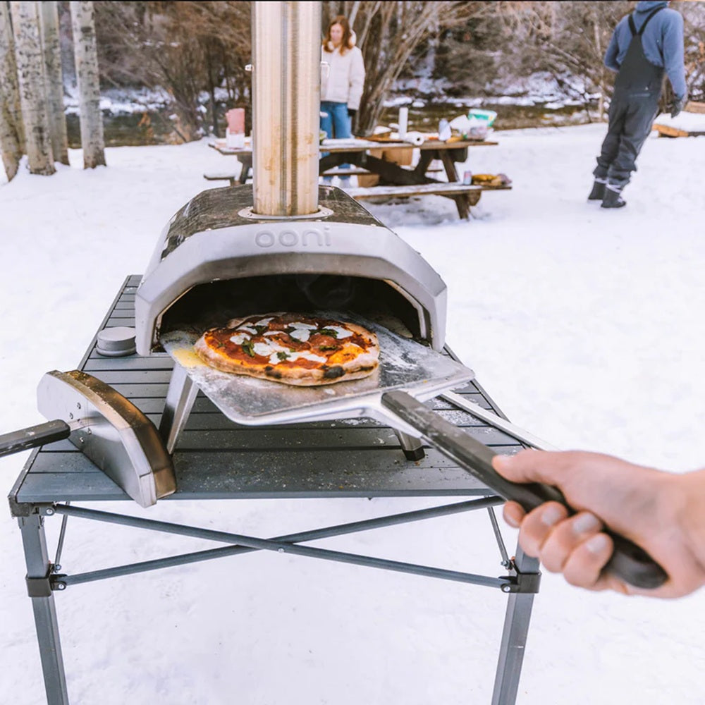 Portable pizza oven being used to cook a pizza in a snowy outdoor setting.