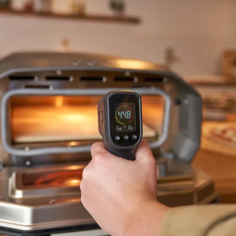Hand holding a digital thermometer in front of an open pizza oven showing a temperature reading.