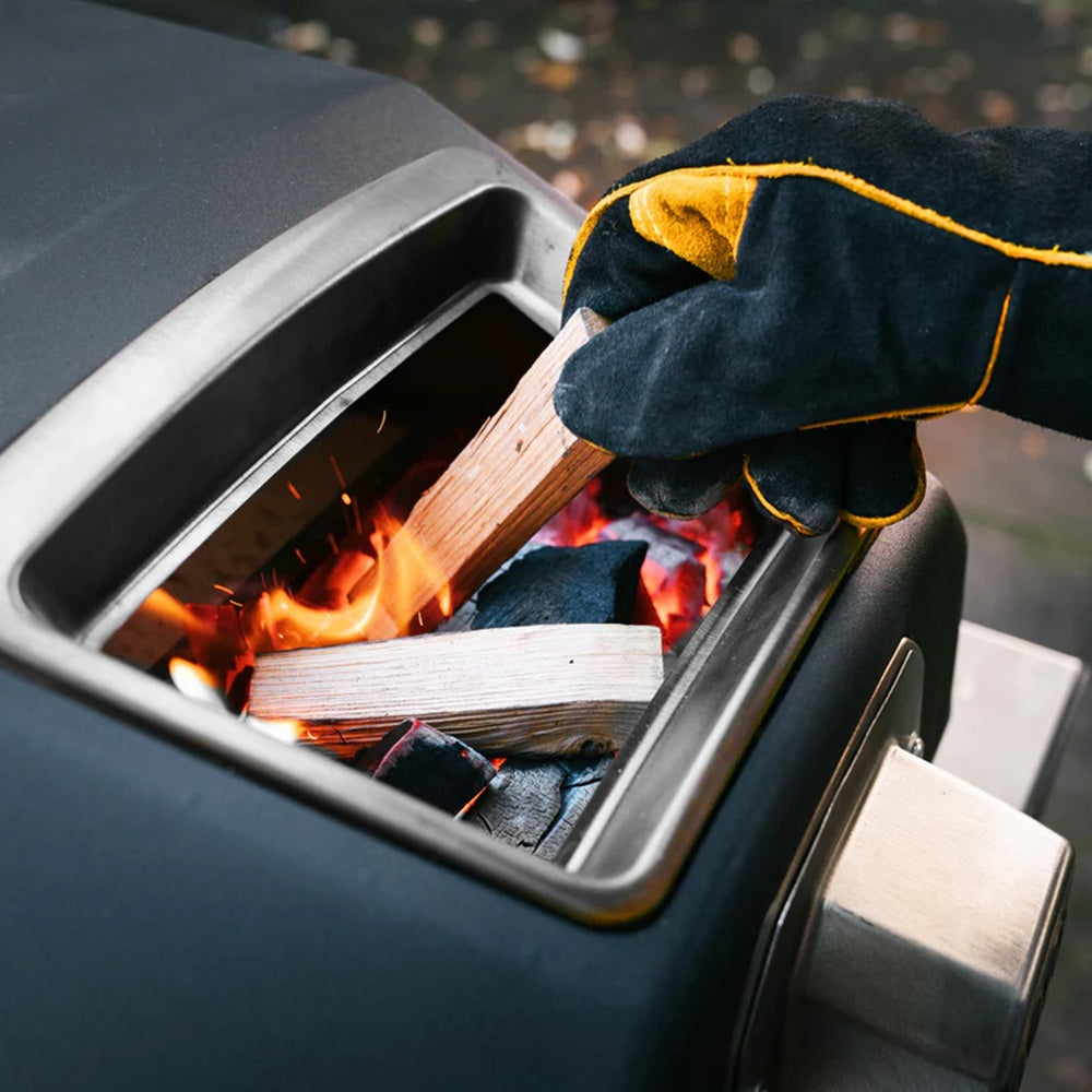 Hand wearing a glove using a wooden stick to light firewood inside a barbecue grill.