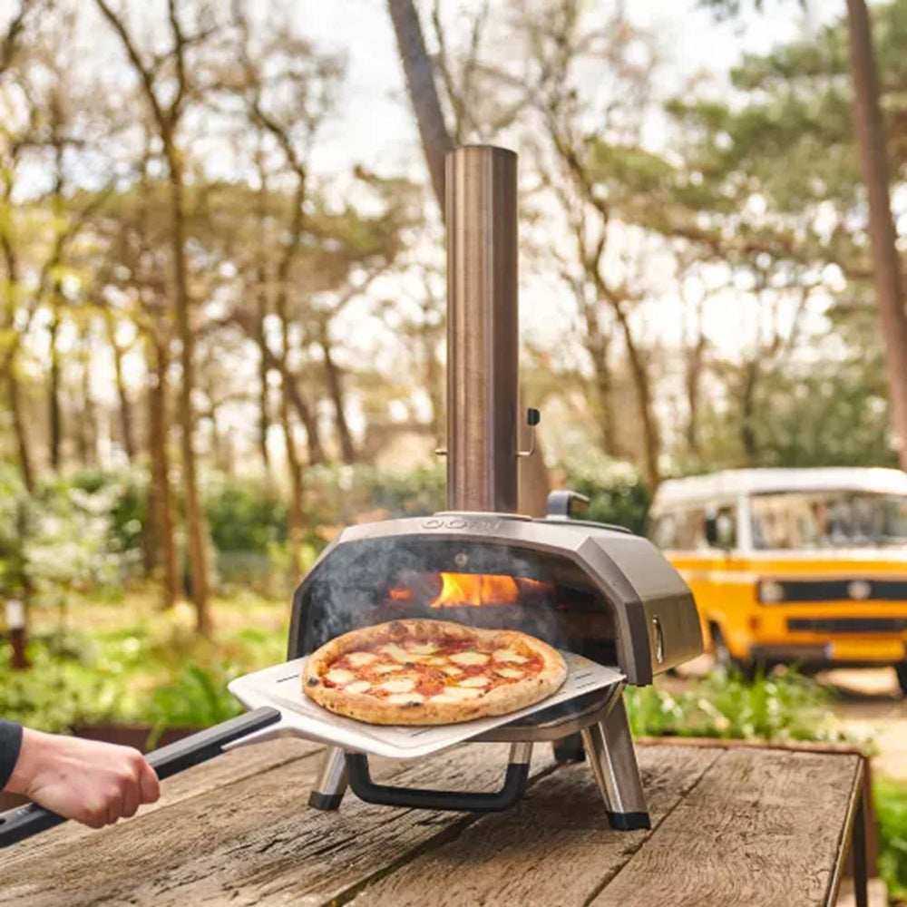 Portable pizza oven with a pizza being cooked outdoors, with a van in the background.