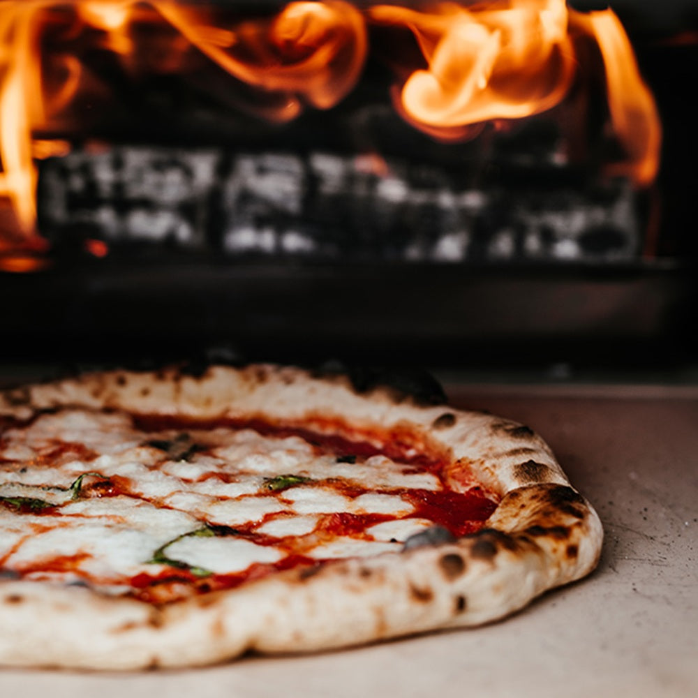 Margherita pizza being cooked in a wood-fired oven with flames.