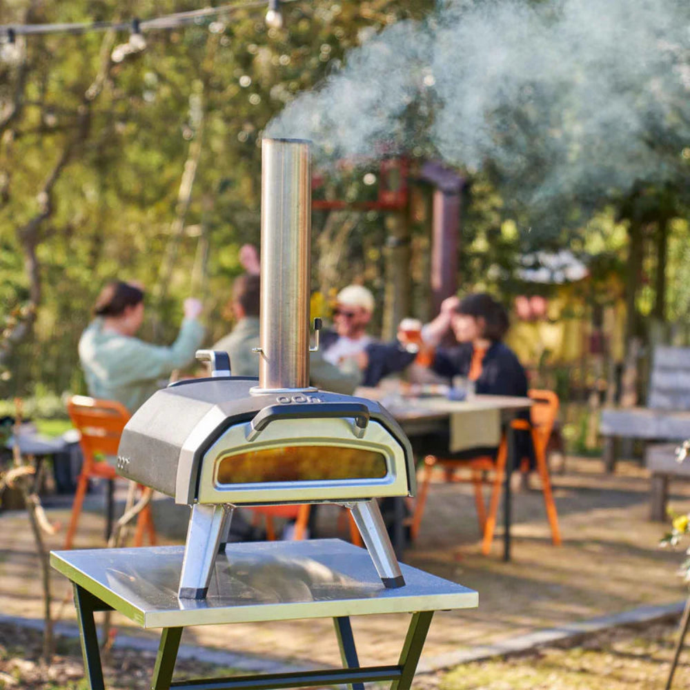 Outdoor pizza oven with smoke rising, people sitting at tables in the background