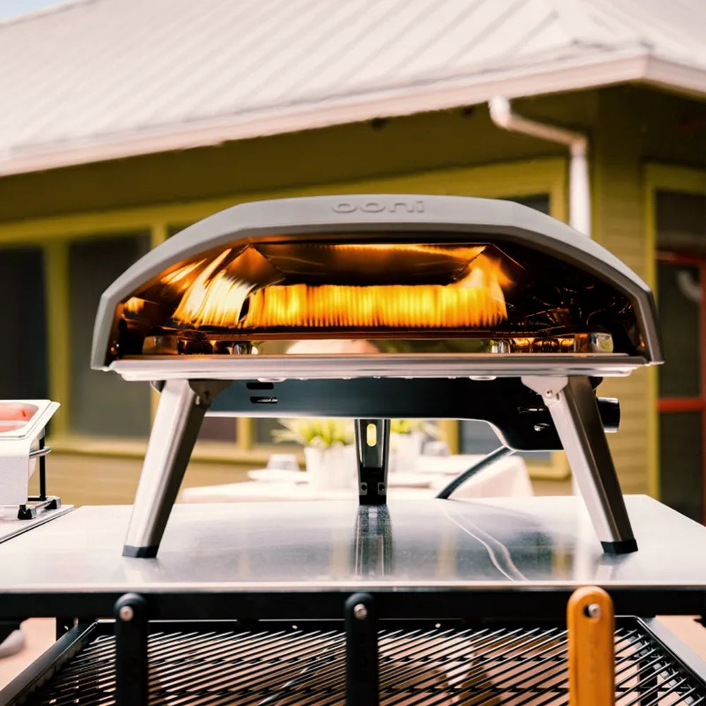 Outdoor pizza oven on a stand with glowing interior, set against a blurred outdoor background.