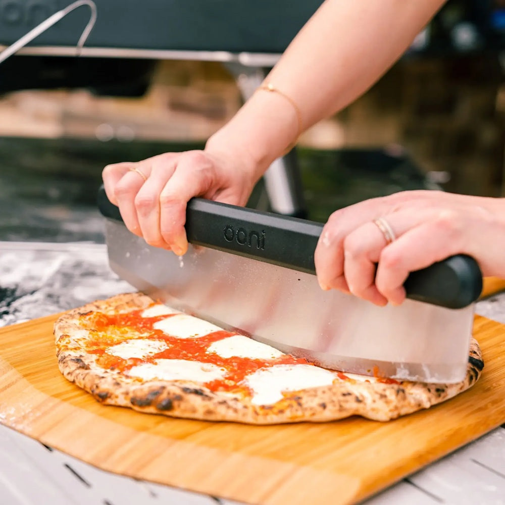 Person cutting a pizza with a knife on a wooden board outdoors