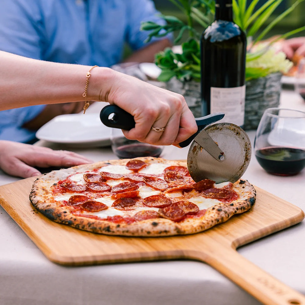 Person cutting a pepperoni pizza on a wooden board with a pizza cutter, surrounded by wine and plants.