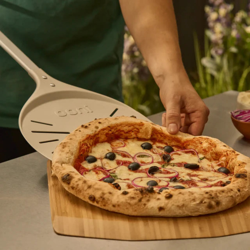 Person using a pizza peel to lift a pizza from a wooden cutting board 