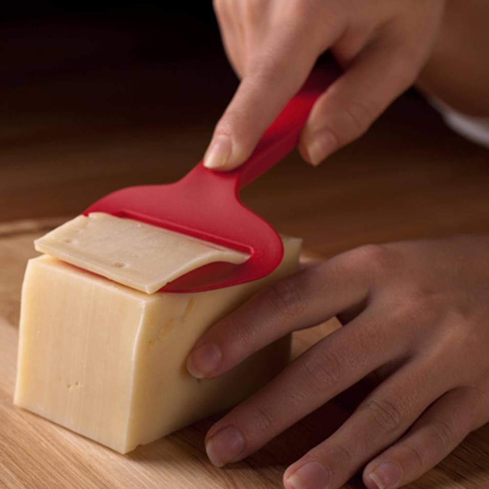 Person using a red cheese slicer to cut a block of cheese on a wooden surface