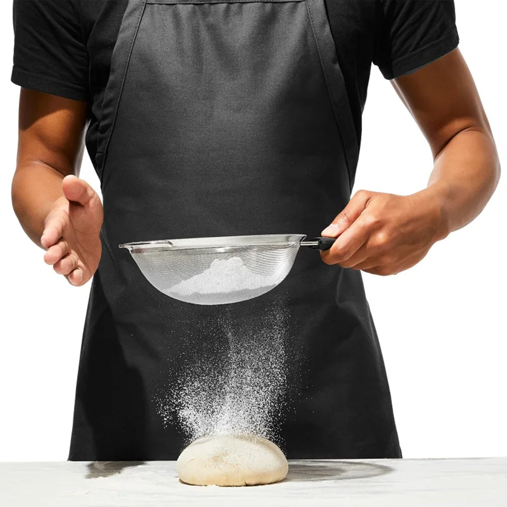 Person sifting flour over a ball of dough with a black apron on a white background