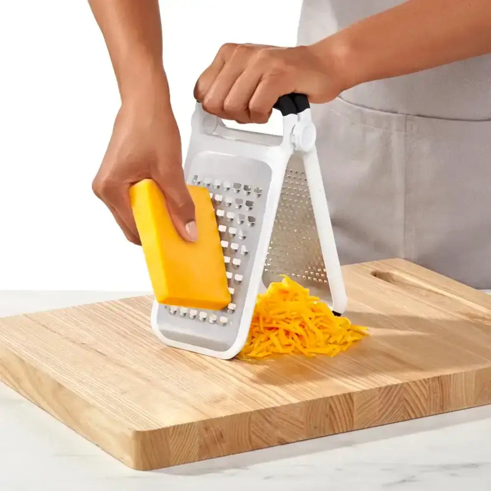Person using a yellow and white grater on a wooden cutting board with shredded yellow vegetable.