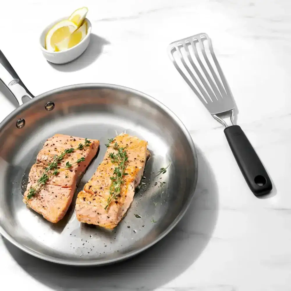 Two pieces of salmon in a frying pan with a spatula and lemon wedge on a white surface